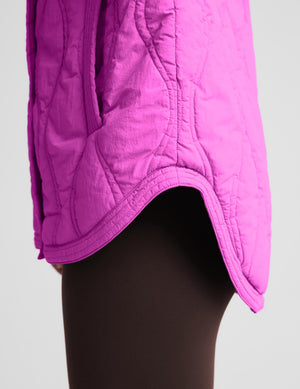 Close-up of a bright pink quilted puffer jacket worn over dark brown tights, showing the curved hem and quilted stitching details against a light gray background.