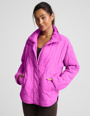 A woman wearing a bright pink quilted zip-up jacket with a stand-up collar and front pockets, photographed against a light gray studio background.