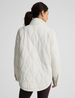 A woman shown from the back wearing a cream-colored quilted jacket with a high stand-up collar, set against a plain light gray studio background.
