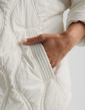 Close-up of a person's hand reaching into a pocket of a cream-colored quilted jacket, showing the texture of the fabric and stitching.