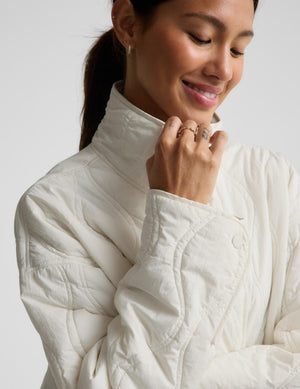 A close-up of a woman wearing a cream-colored quilted jacket, showing the texture of the fabric and the high collar. She is smiling and looking down, with her hand near her chin.