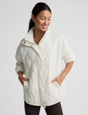 A woman wearing a cream-colored quilted jacket with a stand-up collar and front zipper, standing against a light gray studio background.