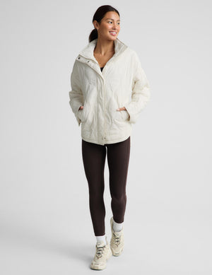 A woman modeling a cream quilted puffer jacket, paired with dark brown leggings and beige sneakers, against a solid light gray background.
