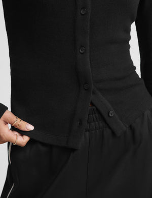 Close-up of a person wearing a black ribbed button-front cardigan and black pants, with a hand lifting the hem of the cardigan to show the elasticized waist detail.