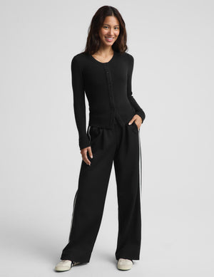 A woman modeling a black long-sleeve button-front top and matching black wide-leg pants with white side stripes, photographed against a light gray studio background.