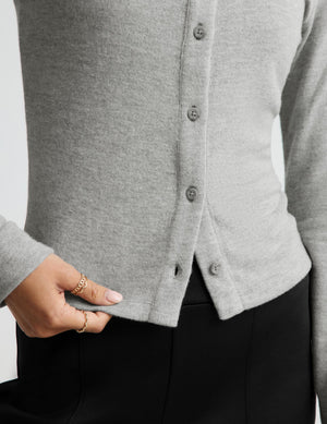 Close-up of a person wearing a light gray knit cardigan with dark buttons, paired with black bottoms. A hand with rings is visible at the side.