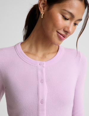 A close-up of a woman wearing a light pink ribbed knit cardigan with a buttoned neckline, shown against a plain light gray background.
