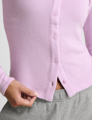 A close-up of a person wearing a light pink ribbed knit cardigan with matching buttons, paired with gray sweatpants. The person's hand is pulling the hem of the cardigan slightly to show the fit.