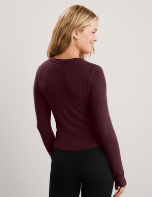A woman wearing a long-sleeve button-front cardigan in deep burgundy, paired with black pants, photographed against a plain light gray studio background.