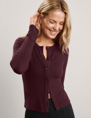 A woman wearing a long-sleeve button-front cardigan in deep burgundy, paired with black pants, photographed against a plain light gray studio background.