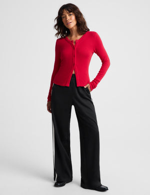 A woman modeling a bright red long-sleeve button-front cardigan with black wide-leg pants featuring white side stripes, photographed against a light gray studio background.