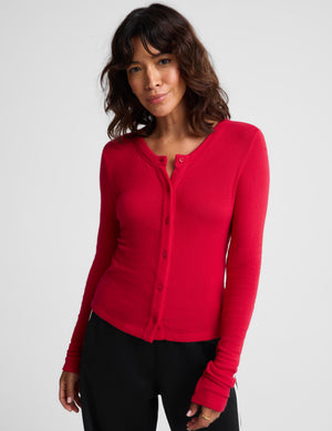 A woman wearing a bright red ribbed long-sleeve button-front cardigan with a split neckline, paired with black pants, photographed against a light gray studio background.