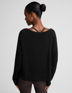 A woman modeling a black long-sleeve scoop-neck top with a relaxed fit, paired with dark brown leggings, against a solid light gray background.