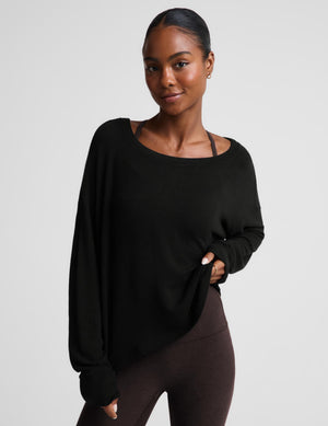 A woman modeling a black long-sleeve scoop-neck top with a relaxed fit, paired with dark brown leggings, against a solid light gray background.