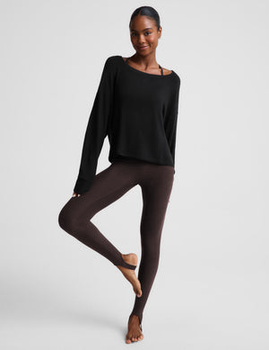 A woman modeling a black long-sleeve scoop-neck top with a relaxed fit, paired with dark brown leggings, against a solid light gray background.