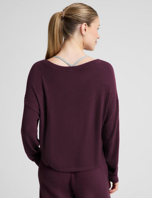 A woman modeling a matching deep plum purple loungewear set, featuring a long-sleeve boat-neck top and coordinating sweatpants, against a solid light gray background.