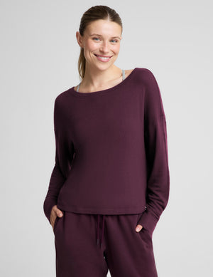 A woman modeling a matching deep plum purple loungewear set, featuring a long-sleeve boat-neck top and coordinating sweatpants, against a solid light gray background.
