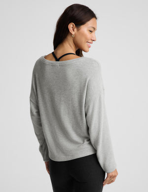 A woman shown from the back wearing a light gray knit sweater with a wide, slightly slouchy neckline and black leggings, photographed against a plain light gray studio background.