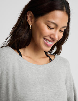 A close-up of a woman wearing a light gray crewneck sweater, showing her smiling and looking down. She has dark brown hair and is wearing a small gold hoop earring.