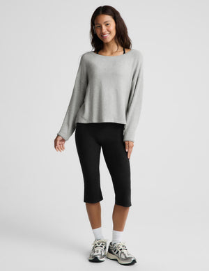 A woman modeling a light gray long-sleeve knit top and black capri leggings against a plain light gray studio background.