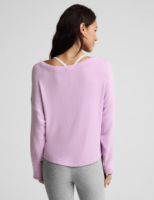 A woman shown from the back wearing a light purple long-sleeve knit top with a wide, open back design that reveals white underwire bra straps, paired with gray leggings, against a light gray studio background.