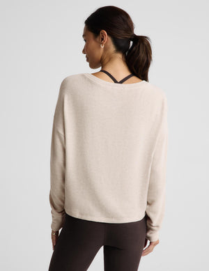 A woman shown from the back wearing a light beige long-sleeve knit top with a black crisscross underband at the neckline, paired with dark brown leggings, against a light gray studio background.