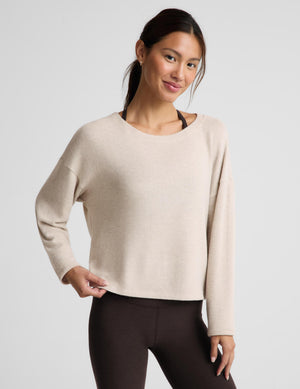 A woman modeling a light beige long-sleeve knit top with a crew neckline, paired with dark brown leggings, against a solid light gray background.