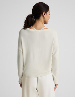 A woman shown from the back wearing a cream-colored knit sweater with a double-strap open neckline and matching cream pants, set against a plain light gray studio background.
