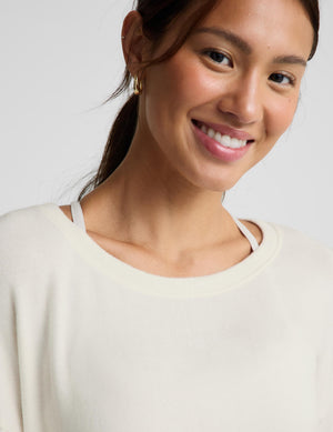 A close-up of a smiling woman wearing a cream-colored crewneck knit top with thin shoulder straps, set against a plain light gray background.