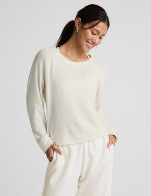 A woman wearing a cream-colored long-sleeve knit top and matching cream sweatpants, standing against a light gray studio background.