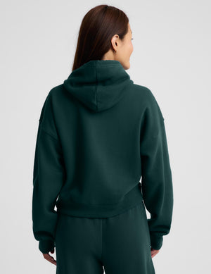 A woman wearing a dark forest green full-zip hoodie and matching sweatpants, standing against a light gray studio background.