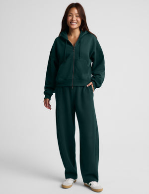 A woman wearing a dark forest green full-zip hoodie and matching sweatpants, standing against a light gray studio background.