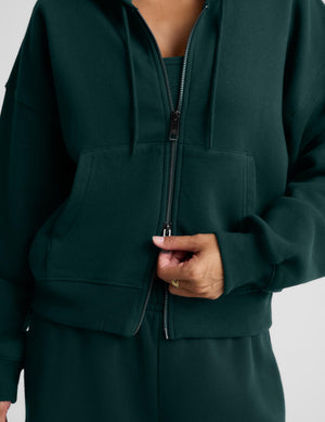 A woman wearing a dark forest green full-zip hoodie and matching sweatpants, standing against a light gray studio background.