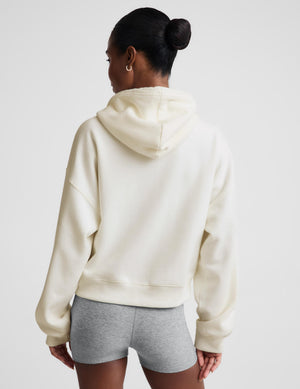A woman modeling a cream-colored zip-up hoodie with a relaxed fit, paired with a gray sports bra and heather gray bike shorts, against a solid light gray background.