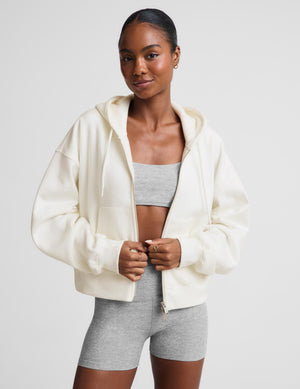 A woman modeling a cream-colored zip-up hoodie with a relaxed fit, paired with a gray sports bra and heather gray bike shorts, against a solid light gray background.