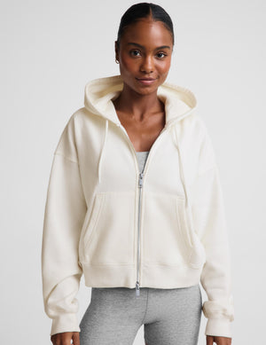 A woman modeling a matching cream-colored lounge set consisting of a zip-up hoodie styled with a gray sports bra and white sneakers against a light gray studio background.