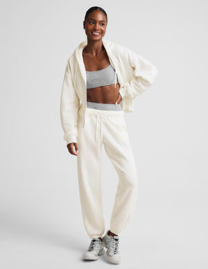 A woman modeling a matching cream-colored lounge set consisting of a zip-up hoodie and jogger pants, styled with a gray sports bra and white sneakers against a light gray studio background.