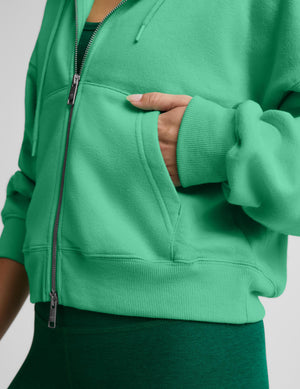 A close-up of a person wearing a bright green zip-up hoodie, showing the front zipper, drawstrings, ribbed cuffs, and a hand tucked into a front pocket.  