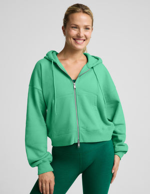 A woman wearing a bright green cropped zip-up hoodie with a drawstring hood and matching green leggings, photographed against a light gray studio background.