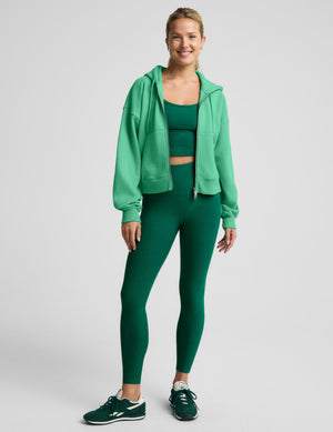 A woman modeling a matching green activewear set consisting of a zip-up hoodie, cropped tank, and leggings, paired with dark green sneakers, against a light gray studio background.