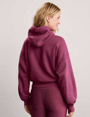 A smiling blonde woman wearing a mauve-red zip-up hoodie and matching leggings against a light gray studio background.