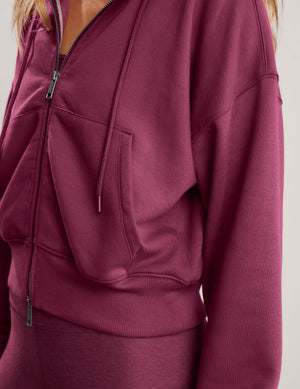 A blonde woman wearing a mauve-red zip-up hoodie and matching leggings against a light gray studio background.