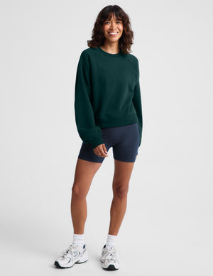 A woman modeling a dark green crewneck sweatshirt and navy blue athletic shorts against a light gray studio background.