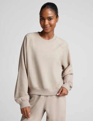 Woman wearing an oatmeal colored crewneck sweatshirt and sweatpants against a plain background. 