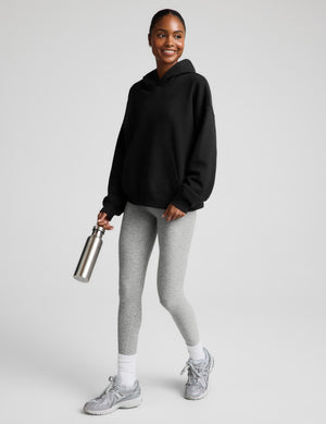 A woman modeling a black pullover hoodie with a front kangaroo pocket, paired with light gray leggings, against a solid light gray background.