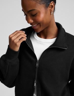 A woman wearing a black cropped zip-up sweatshirt with a stand collar over a white top and black leggings, photographed against a plain light gray studio background.