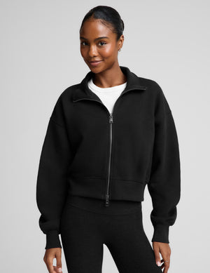 A woman wearing a black cropped zip-up sweatshirt with a stand collar over a white top and black leggings, photographed against a plain light gray studio background.