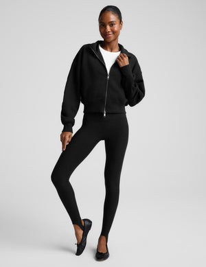A woman wearing a black cropped zip-up sweatshirt with a stand collar over a white top and black leggings, photographed against a plain light gray studio background.