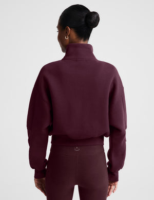 A woman wearing a cropped, zip-up fleece jacket in a deep burgundy color, styled over a white t-shirt and matching burgundy leggings, against a light gray studio background.