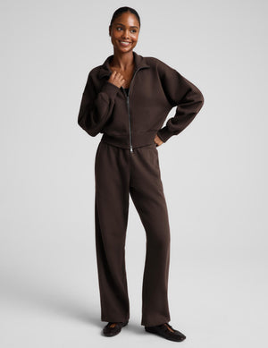 A woman wearing a dark brown zip-up sweatshirt and matching sweatpants against a light gray studio background.